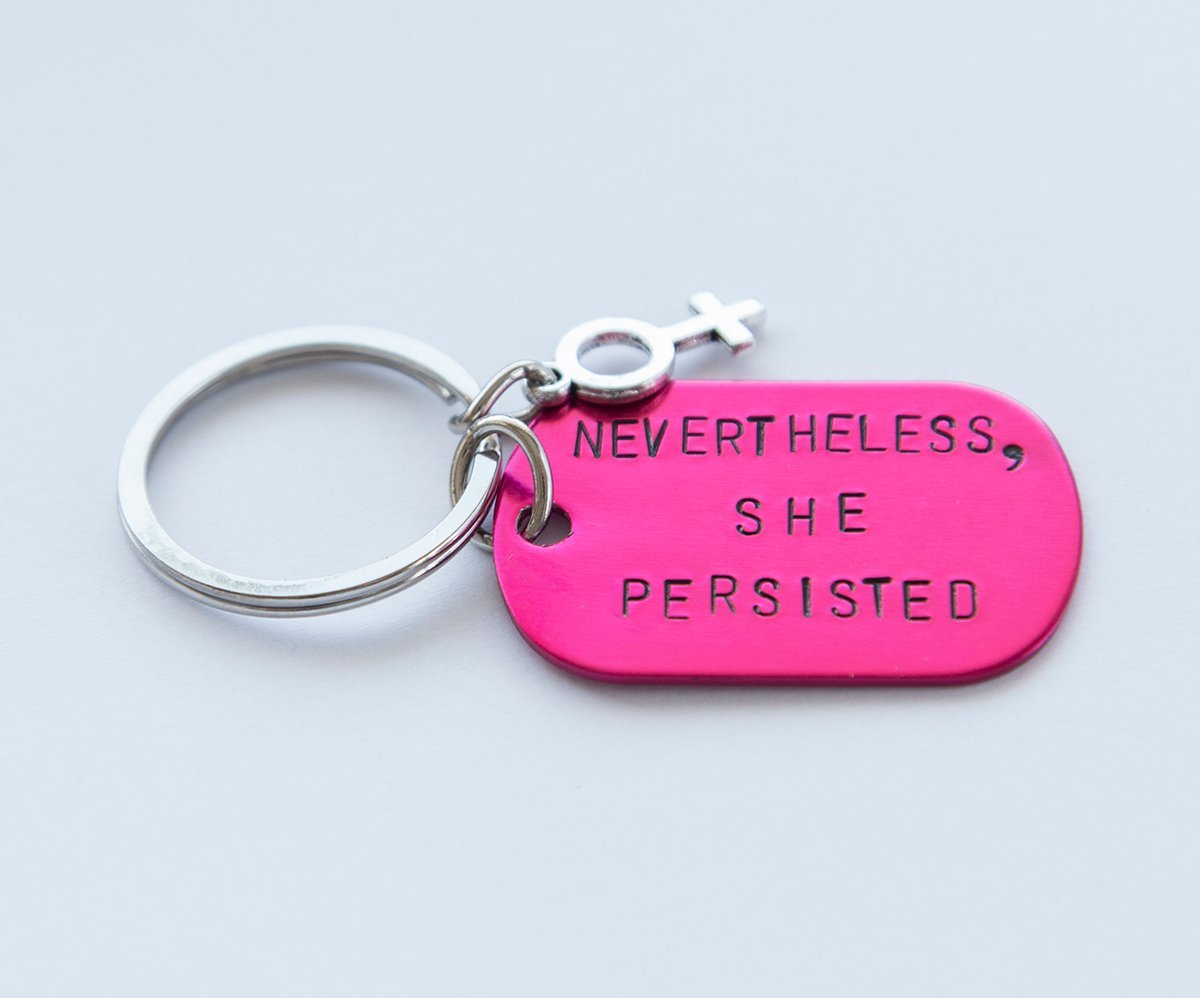 A pink feminist keychain with the phrase 'Nevertheless She Persisted' hand-stamped on a lightweight aluminum tag, attached to a durable stainless steel ring.